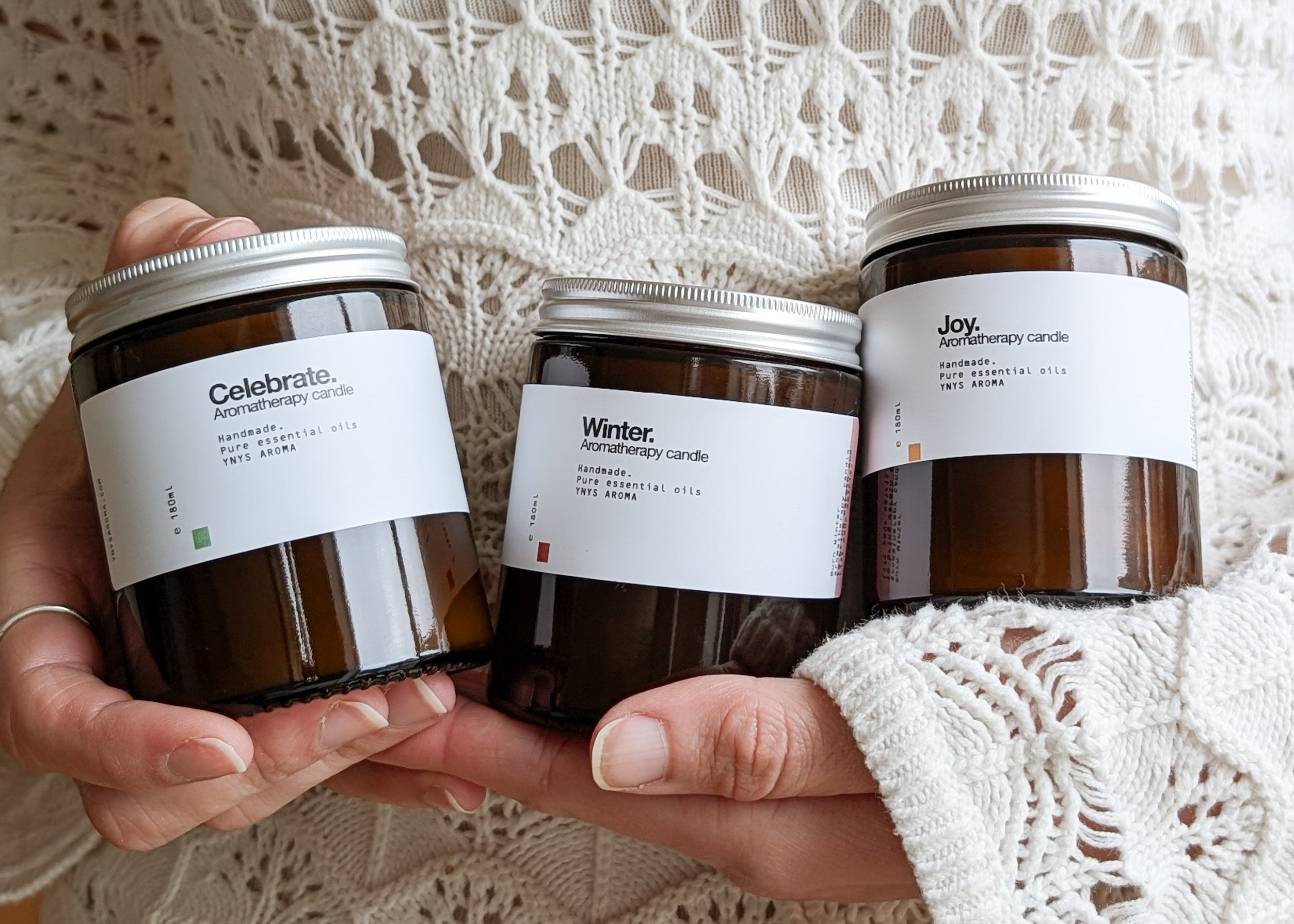 Three aromatherapy candles with labels 'Celebrate', 'Winter' and 'Joy' held by a person wearing a white jumper.